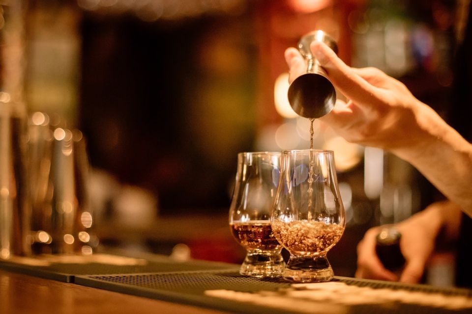 A bartender pours a dark spirit from a metal jigger into two tasting glasses on a bar counter with a warm, dim light.