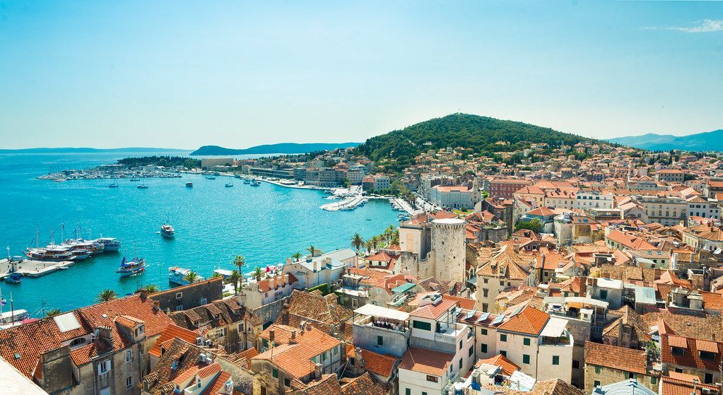 Una vista aerea di una città costiera con tetti in terracotta che si affaccia su un porto blu brillante pieno di barche in una giornata di sole.