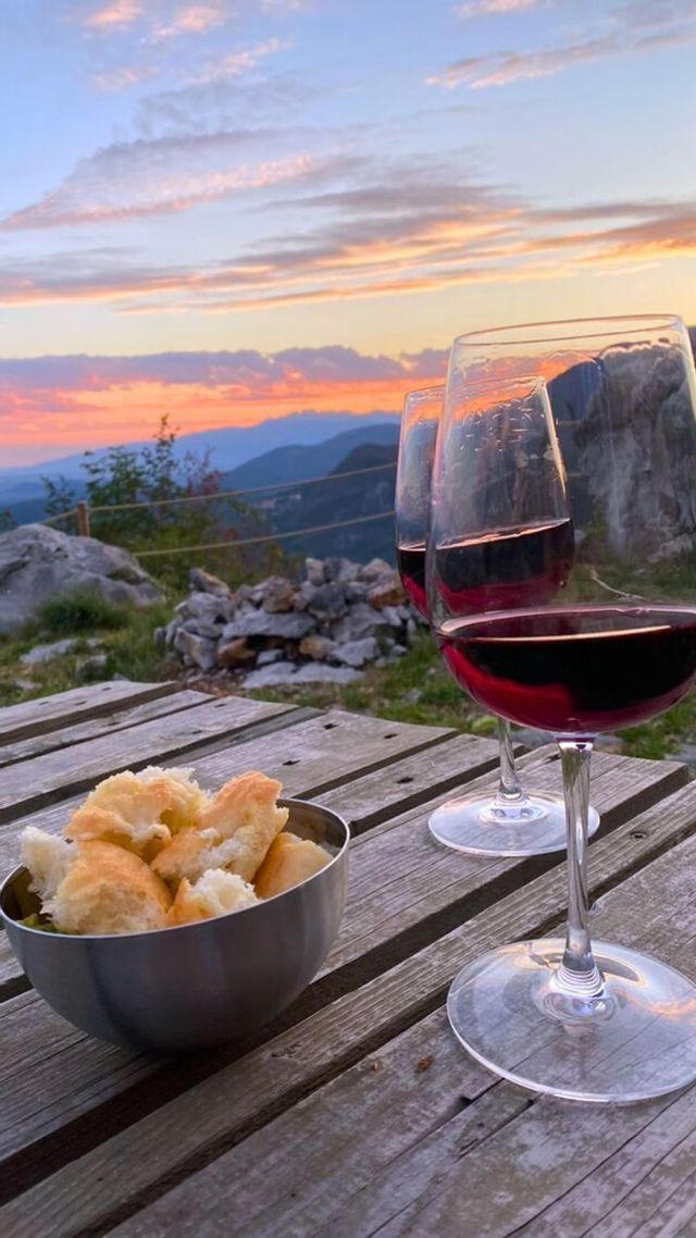 Due calici di vino rosso e una ciotola di pane su un tavolo di legno, con vista sulle montagne al tramonto.