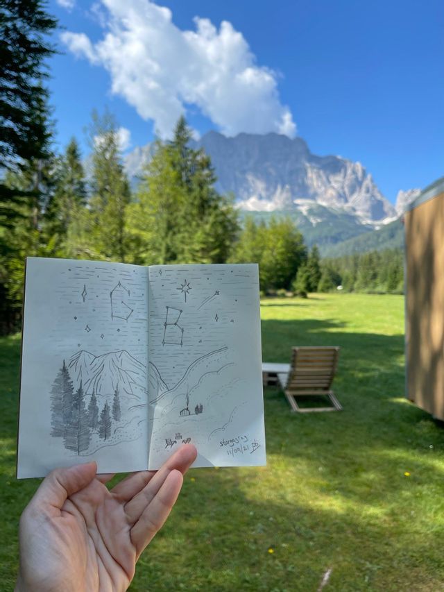 Una mano tiene un quaderno da disegno con uno schizzo a matita di un paesaggio montano notturno, sullo sfondo dell'attuale vista soleggiata delle montagne.