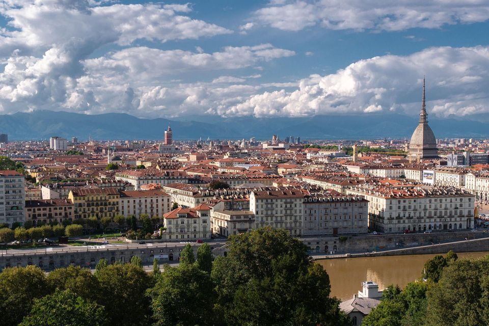 Una vista panorámica de una ciudad con tejados de terracota y una aguja prominente, con montañas a lo lejos bajo un cielo azul nublado.