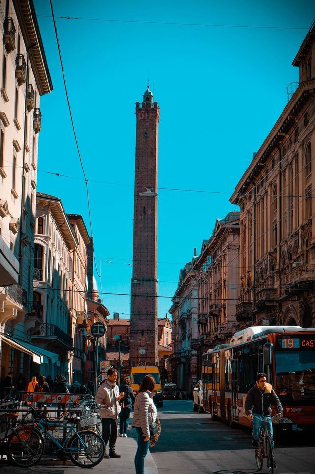 Una alta torre de ladrillo se alza al final de una estrecha calle de la ciudad llena de peatones, bicicletas y un autobús bajo un cielo azul claro.