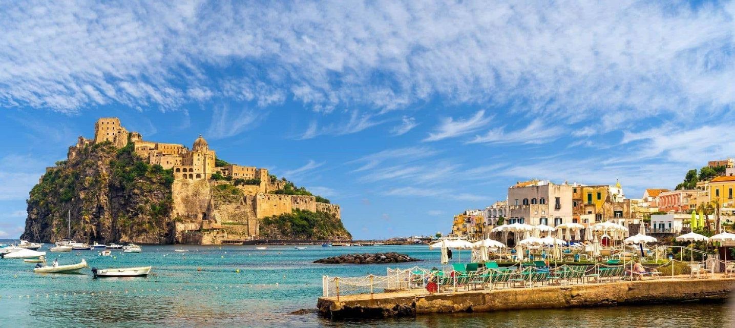Un castello si erge su una scogliera rocciosa che domina una città costiera e una baia piena di barche sotto un cielo azzurro.