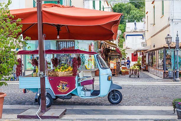 Un camioncino a tre ruote azzurro funge da bancarella di frutta con un grande ombrellone rosso su una strada di ciottoli.
