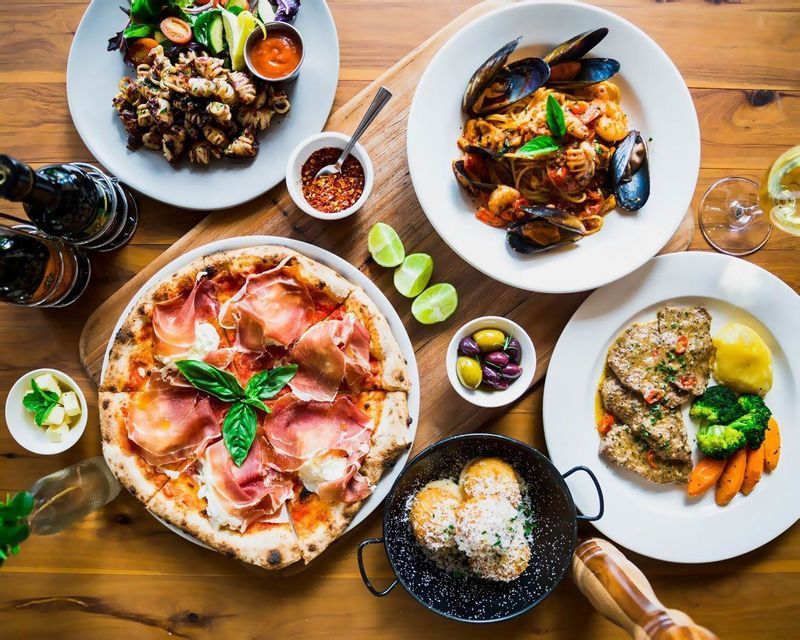 Una vista dall'alto di un tavolo di legno imbandito con vari piatti italiani, tra cui pizza al prosciutto, pasta con le cozze e calamari alla griglia.