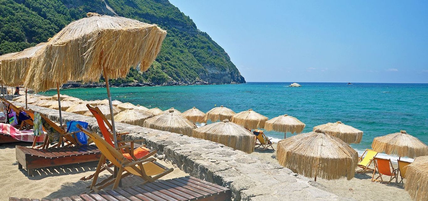 Ombrelloni di paglia e lettini allineati lungo un muro di pietra accanto al mare turchese e a una lussureggiante scogliera verde.