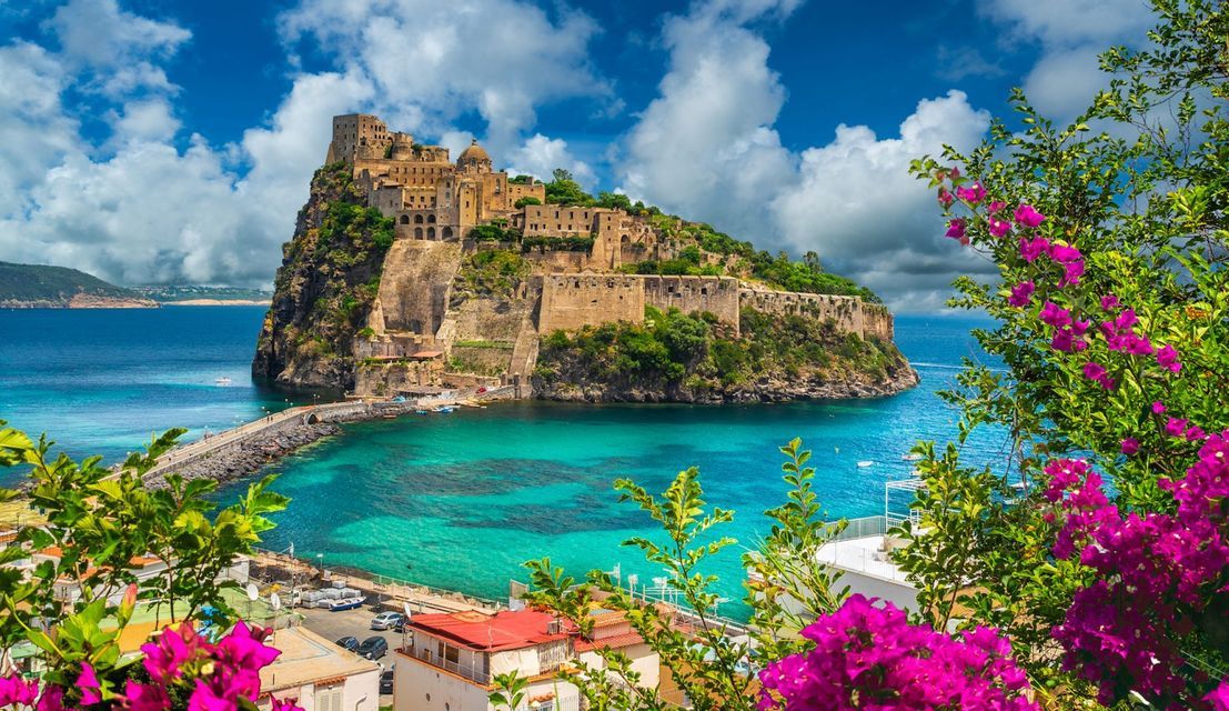 Un castello su un'isola rocciosa si erge in acque turchesi sotto un cielo azzurro nuvoloso, incorniciato da fiori rosa brillanti in primo piano.
