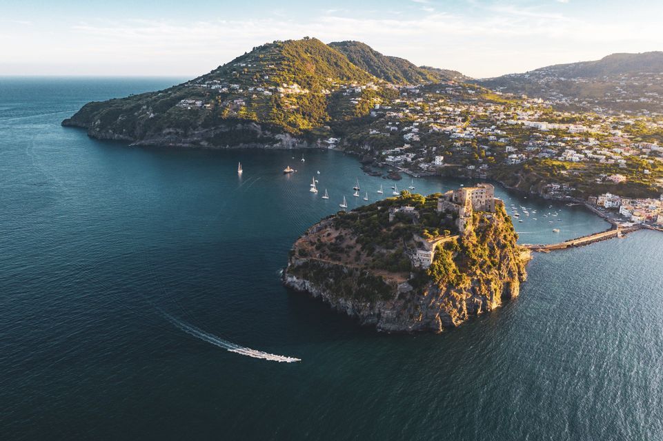 Veduta aerea di un castello su un isolotto roccioso collegato da un ponte a una città sulla terraferma, con barche a vela nella baia.