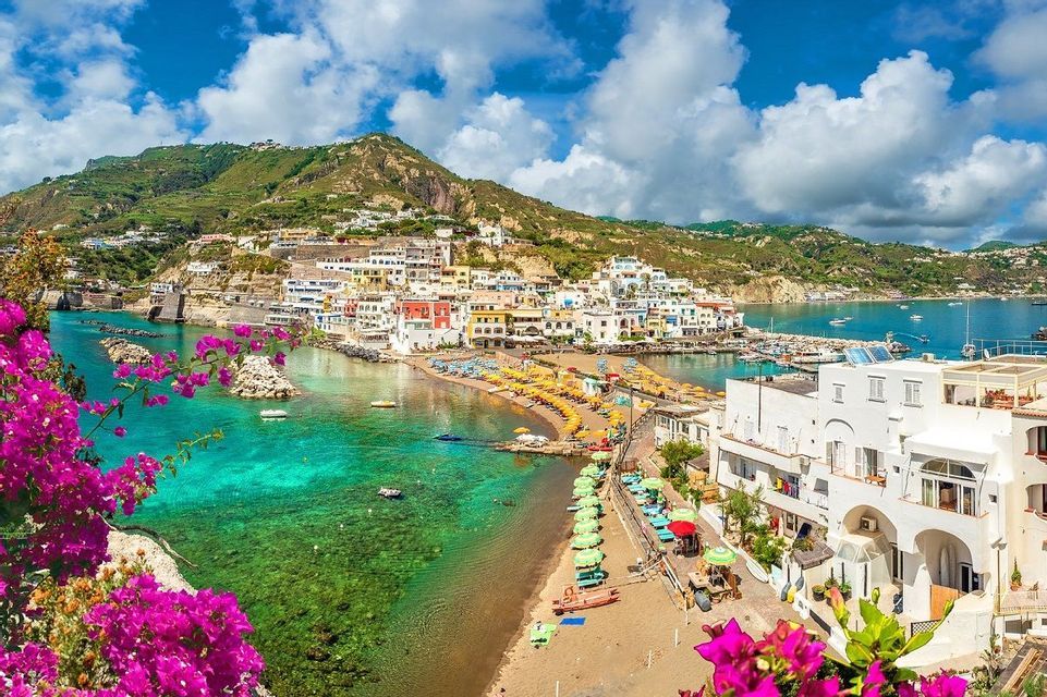Una colorata città costiera si curva attorno a una baia con una spiaggia piena di ombrelloni, acqua turchese e una montagna verde.