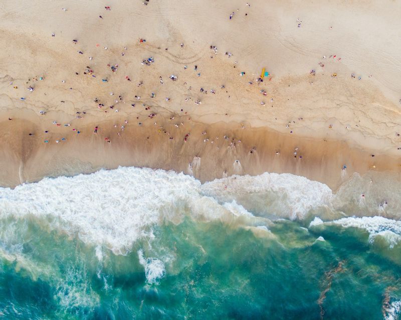 Una vista aerea dall'alto di una spiaggia sabbiosa affollata con persone, mentre onde turchesi dell'oceano con schiuma bianca si infrangono a riva.