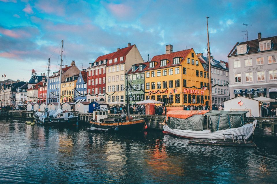 Colorful buildings and boats line a waterfront canal, with their reflections in the water under a pastel-colored sky.