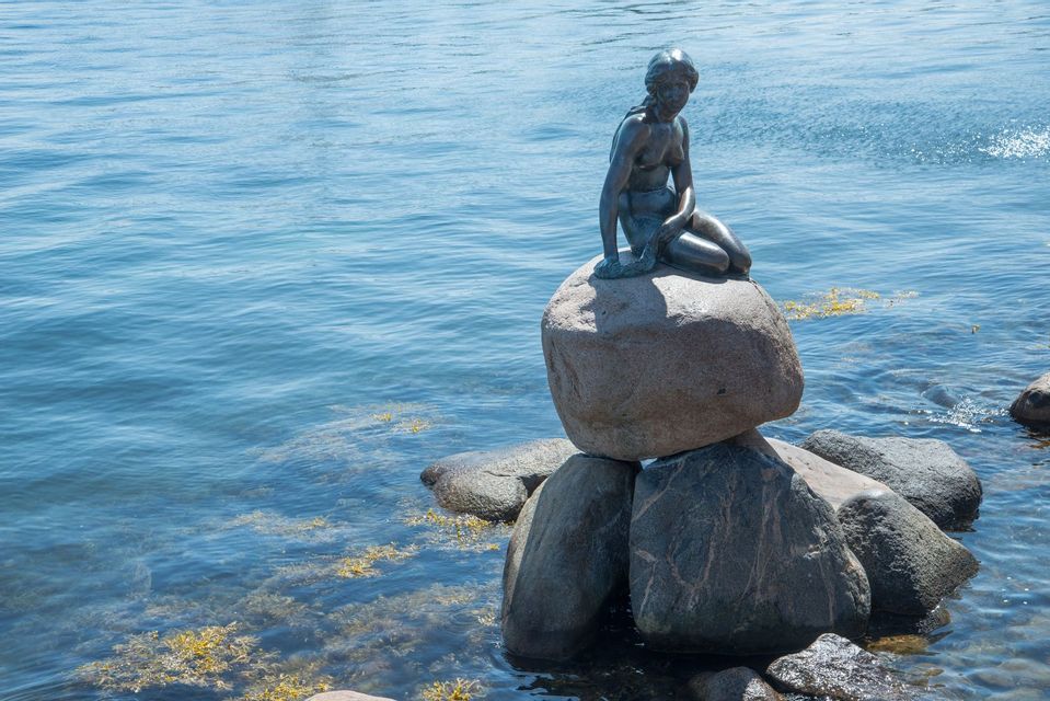 A bronze statue of a mermaid sits on a large rock surrounded by calm blue water.