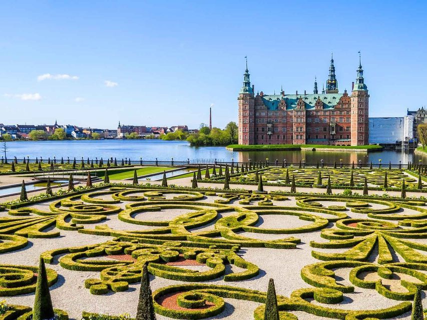 A large, ornate brick castle with a green roof stands beside a lake, with manicured baroque gardens in the foreground on a sunny day.