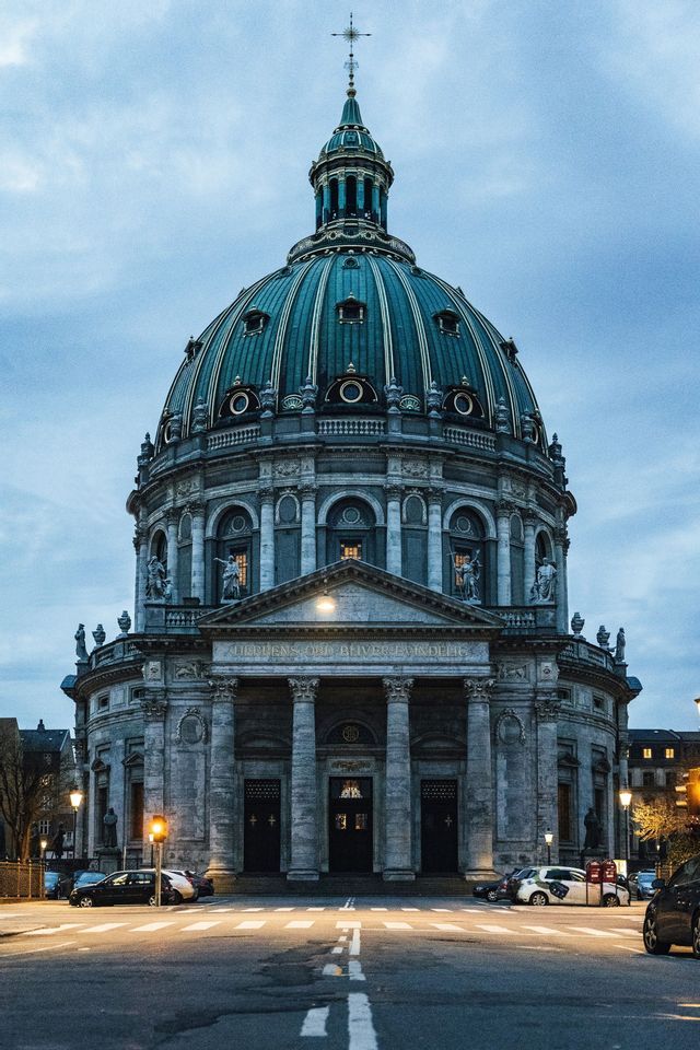 A large stone church with a prominent green dome and classical columns, seen from across a street at dusk.