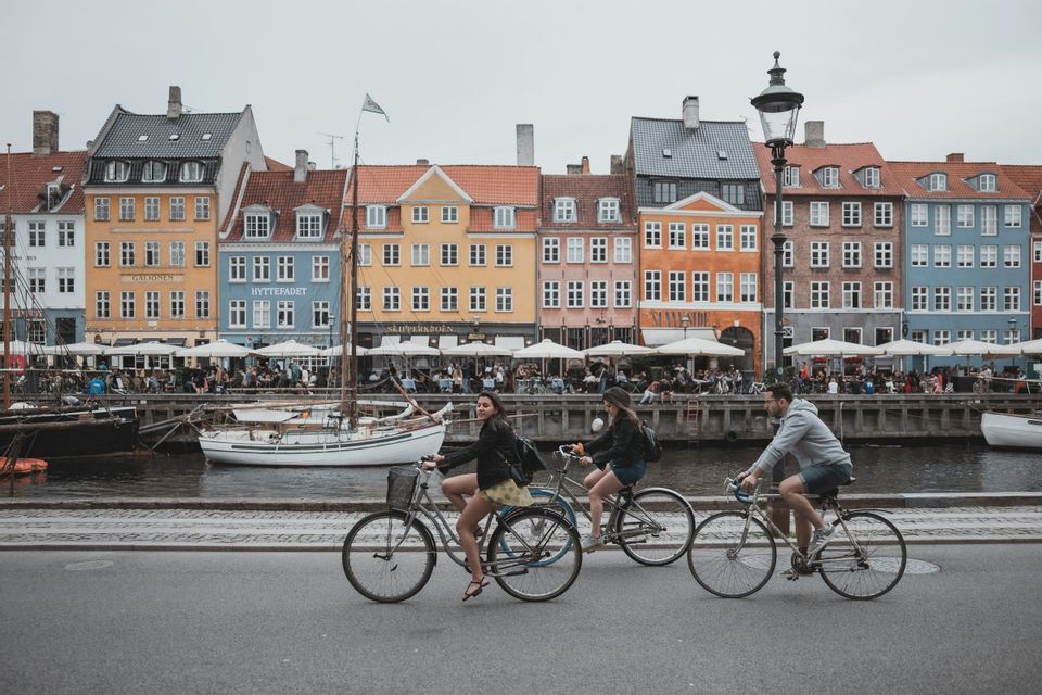 A WeRoad group trip of three people riding bicycles along a canal with colorful waterfront buildings and boats in the background.