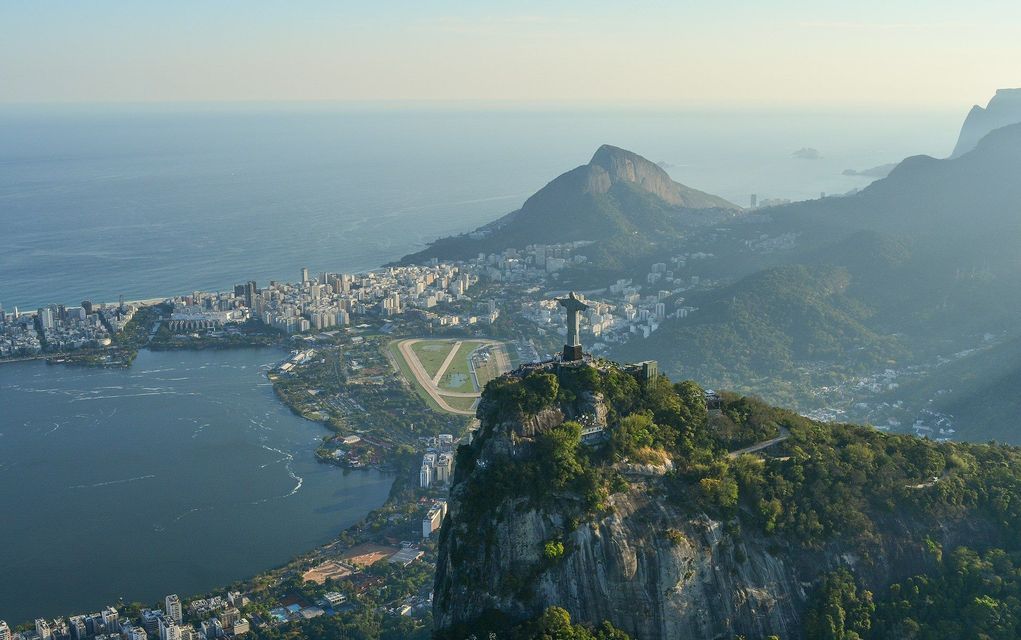Aerial view of the Christ the Redeemer statue on a mountain, overlooking a coastal city with a lagoon and mountains.