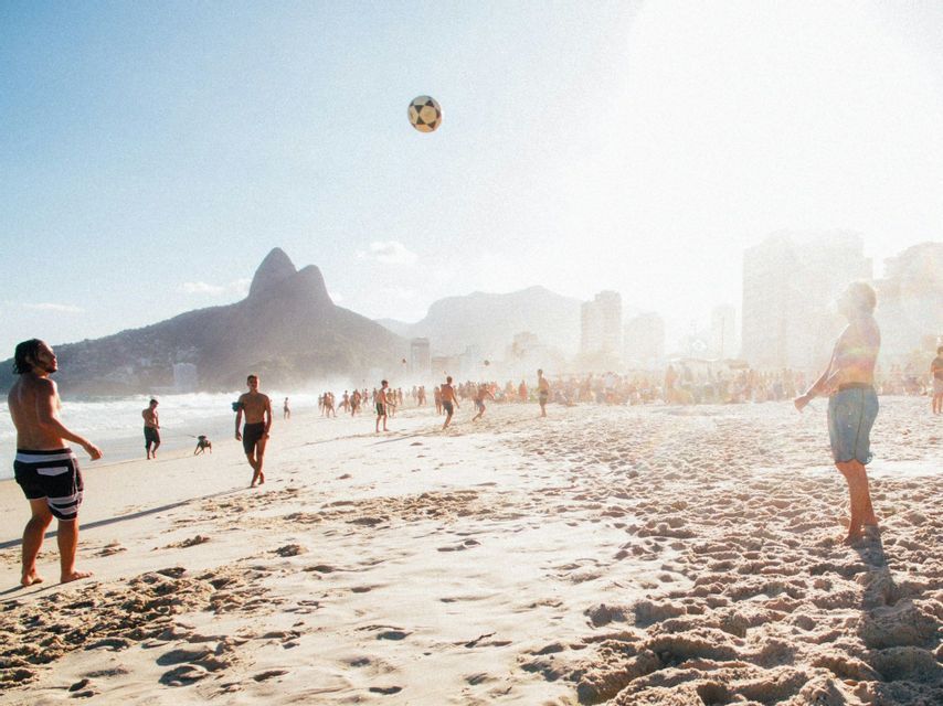 A group of people playing soccer on a crowded, sandy beach with a ball in mid-air and mountains in the background.