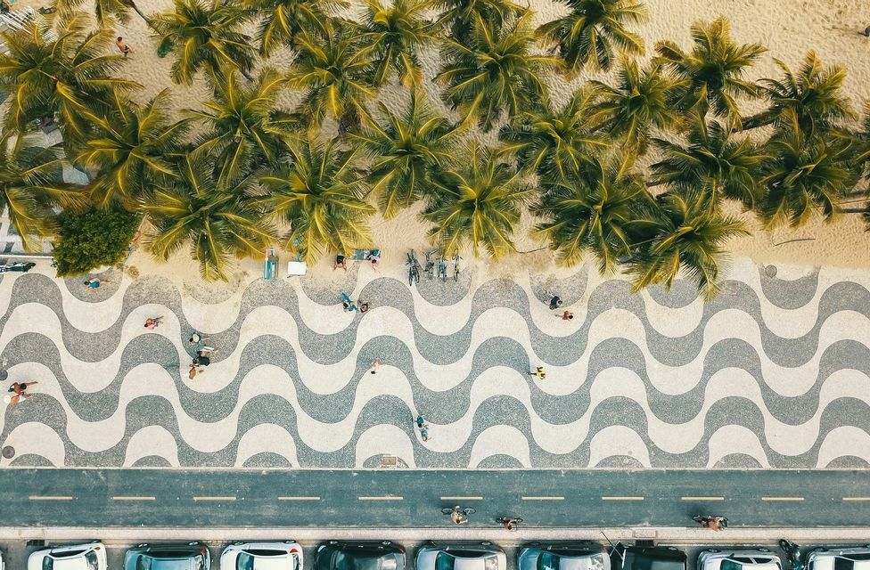 An aerial, top-down view of a beach with palm trees, a promenade with a black and white wavy mosaic pattern, and a road with cars.