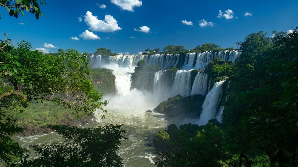 A wide view of a large waterfall system cascading into a river, surrounded by lush green forest under a blue sky with clouds.