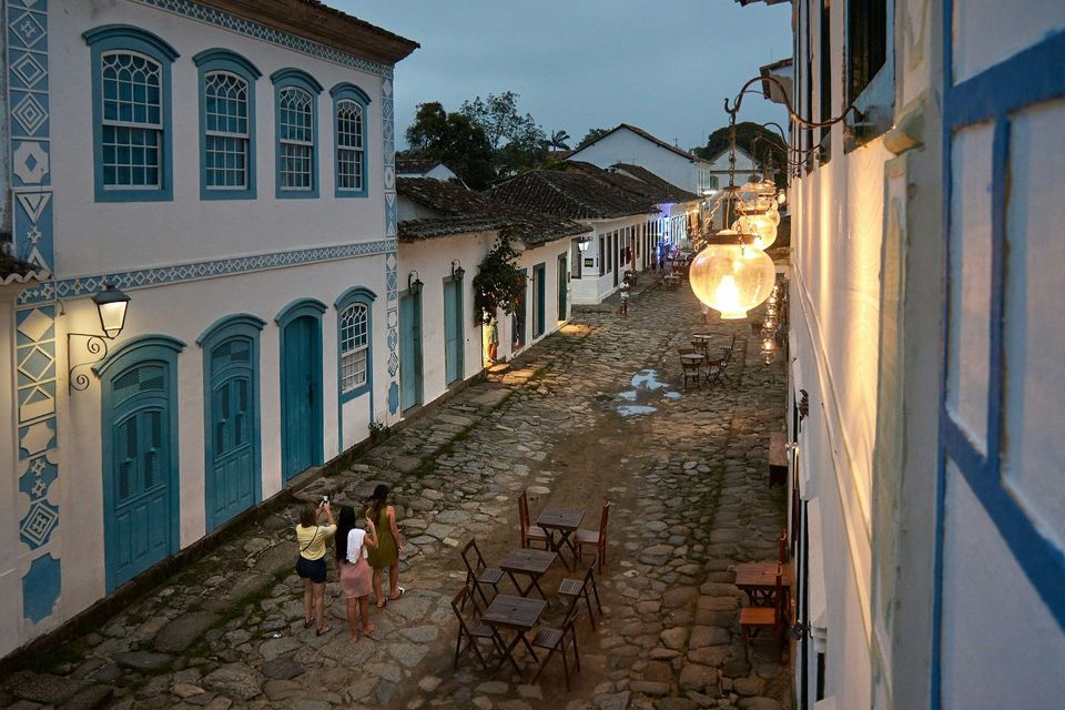 A WeRoad group trip of three women taking photos on a cobblestone street at dusk, lined with colonial buildings and lit lanterns.