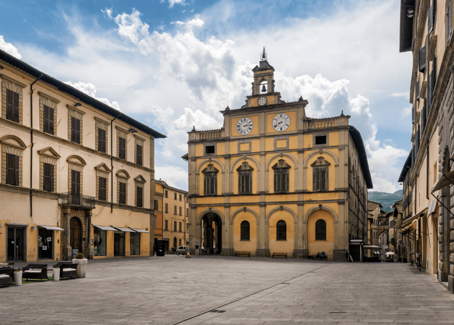 Un grande edificio giallo con una torre dell'orologio e finestre ad arco si erge in una piazza di pietra vuota sotto un cielo nuvoloso.