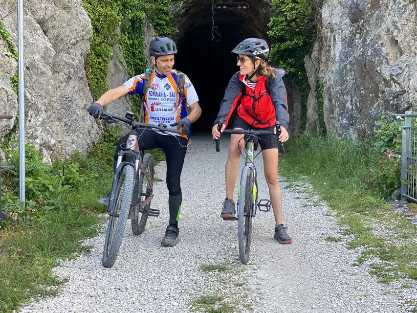 Un uomo e una donna in tenuta da ciclisti stanno con le loro biciclette su un sentiero di ghiaia, parlando davanti all'ingresso di una galleria.