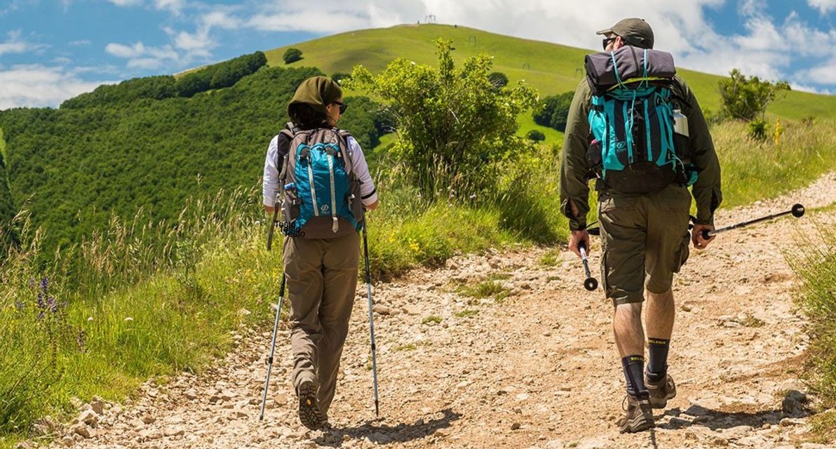 Due persone in un viaggio di gruppo WeRoad che fanno trekking su un sentiero sterrato, attraversando verdi colline, con zaini e bastoncini.