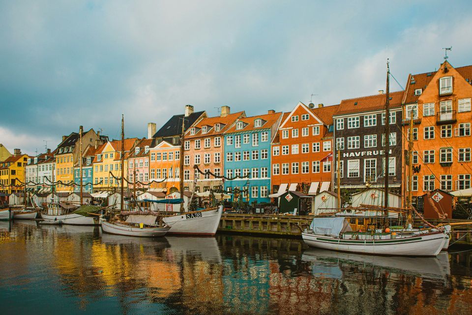 A row of colorful buildings and docked sailboats reflected in a canal under a cloudy sky.