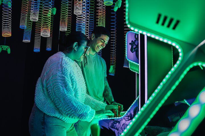 A man and a woman smile while playing a classic arcade game, bathed in green light under hanging colorful spring toys.