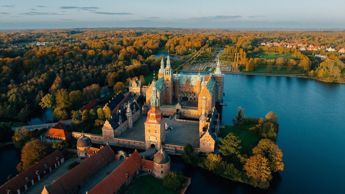 An aerial view of a large castle with spires on a lake, surrounded by a dense forest with autumn colors at sunrise.