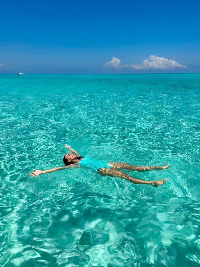 Eine Frau im blauen Badeanzug schwimmt mit ausgestreckten Armen auf dem Rücken im klaren türkisfarbenen Wasser unter einem strahlend blauen Himmel.