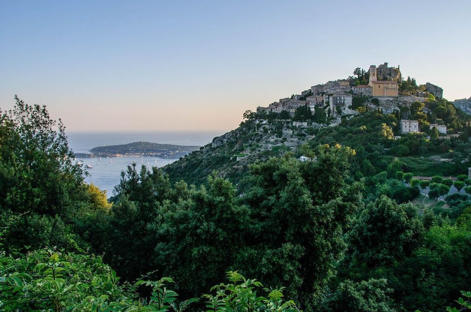 Un pueblo de piedra en una exuberante ladera verde con vistas al mar y una península distante con barcos al atardecer.