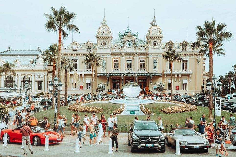 Una multitud de personas se reúne en una plaza con coches de lujo frente a un edificio ornamentado con palmeras.