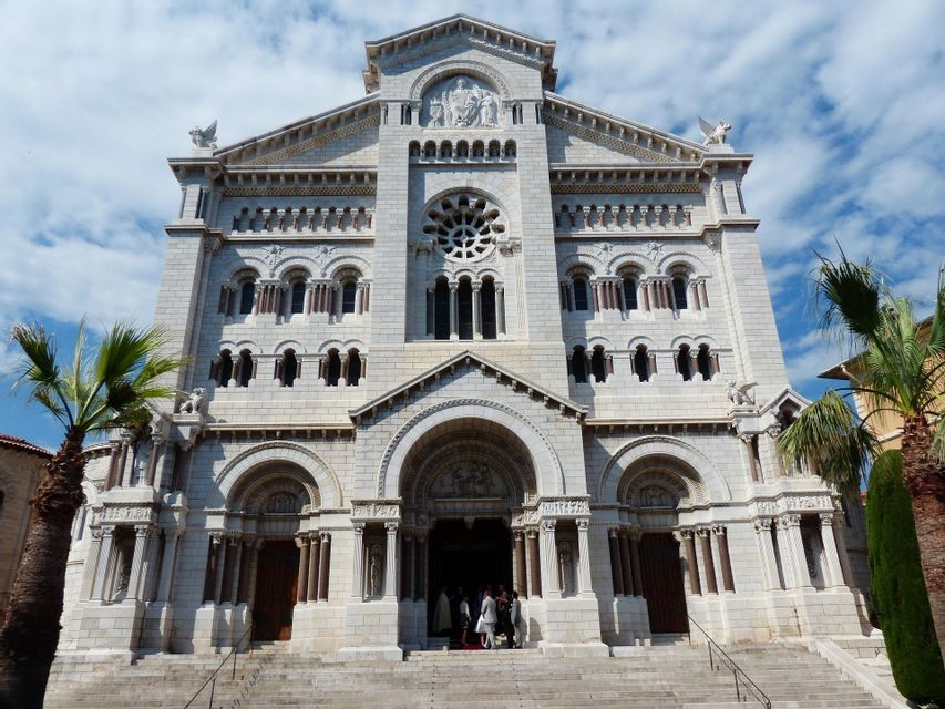 La fachada frontal de una ornamentada catedral de piedra blanca con arcos y un rosetón, vista desde debajo de sus escalones en un día soleado.