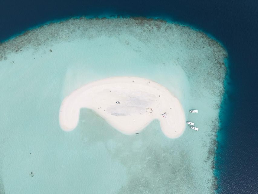 Vue aérienne d'un banc de sable blanc dans des eaux océaniques turquoises, avec des gens se relaxant et des bateaux amarrés à proximité.