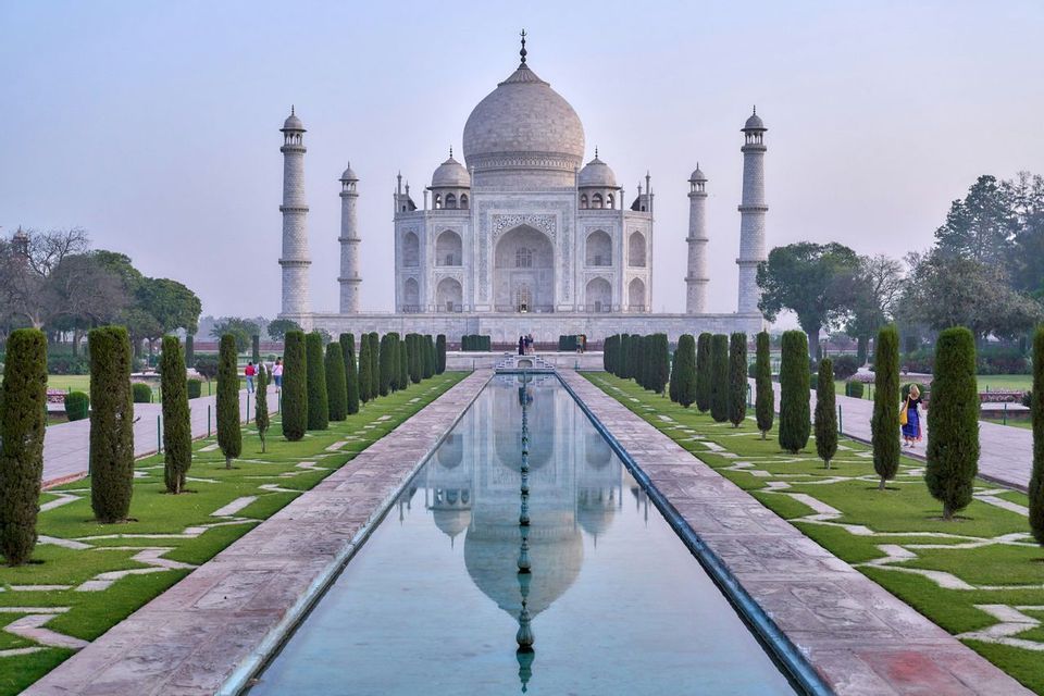 El Taj Mahal se refleja en un largo estanque de agua, flanqueado por jardines bien cuidados con cipreses bajo un cielo pálido.