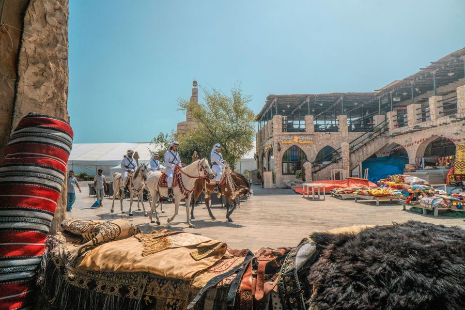Quattro uomini in abiti tradizionali bianchi cavalcano cavalli attraverso una piazza del mercato con edifici in pietra e un minareto.