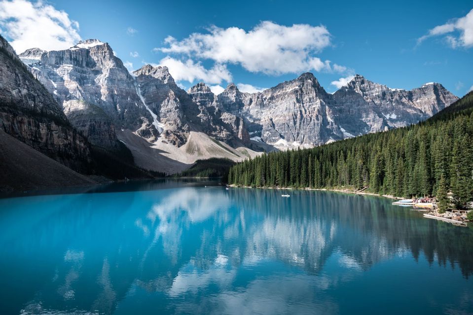 Ein leuchtend blauer Alpensee spiegelt schneebedeckte Berge und einen bewölkten Himmel wider, mit einem dichten Kiefernwald am Ufer.
