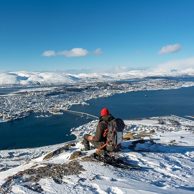 A person with a backpack sits on a snowy mountain peak, looking out at a coastal city and bridge below under a clear blue sky.