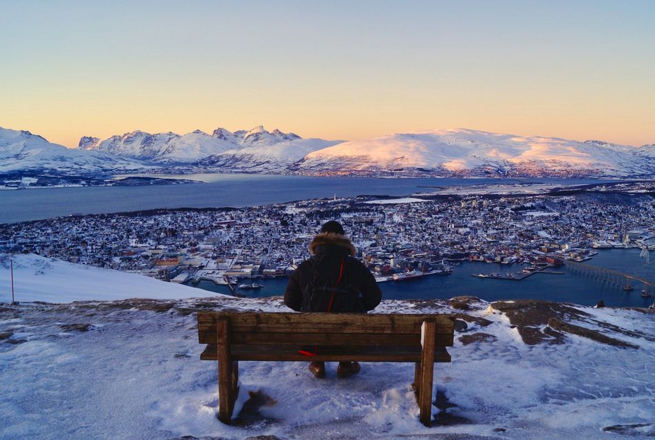 A person sits on a bench on a snowy mountaintop, overlooking a coastal city and distant mountains at sunset.