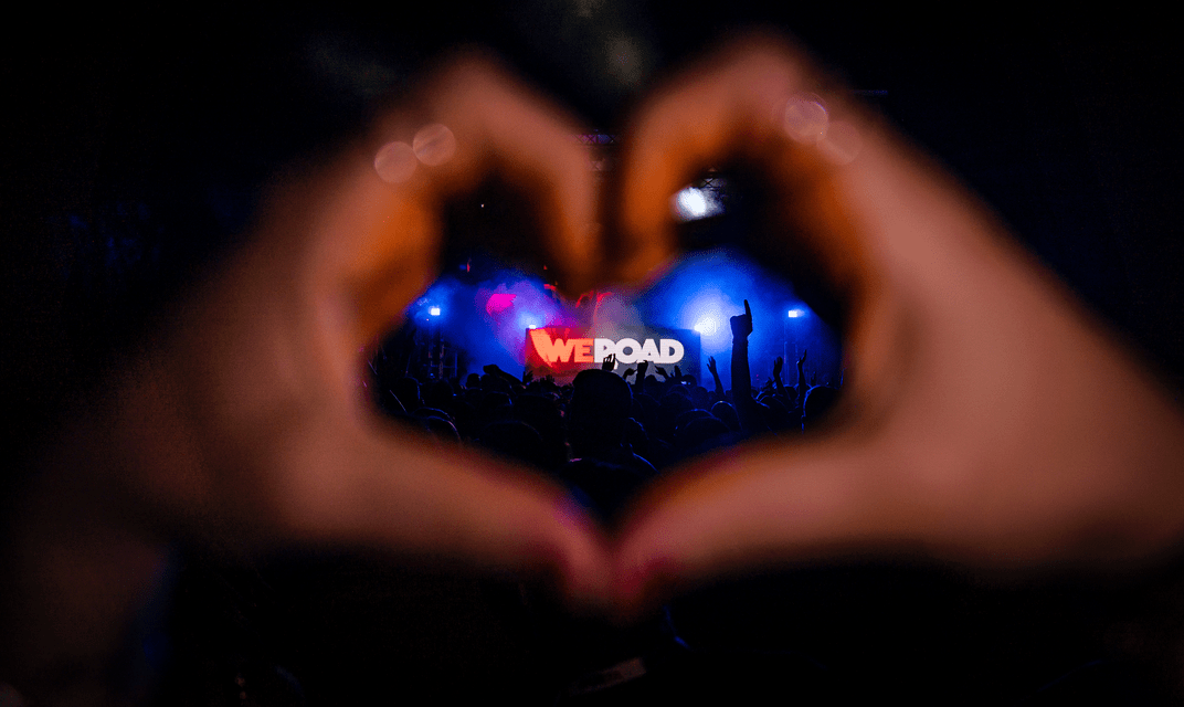 Mani che formano un cuore incorniciano un palco da concerto con una folla e il logo WeRoad su uno schermo.