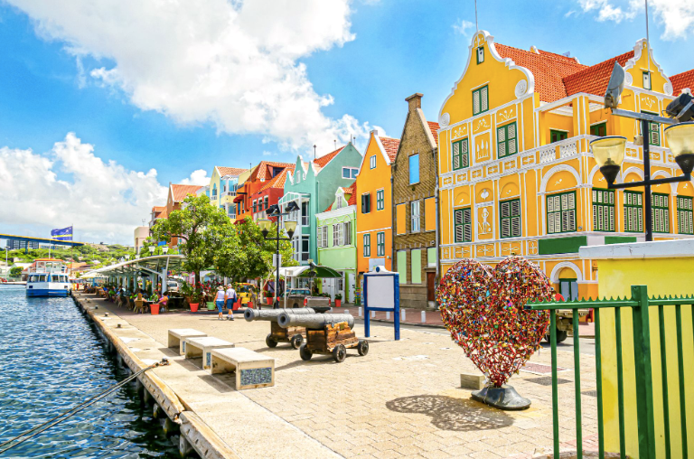 Una passeggiata lungomare con edifici colorati, due cannoni e una scultura a forma di lucchetto a cuore sotto un cielo azzurro.
