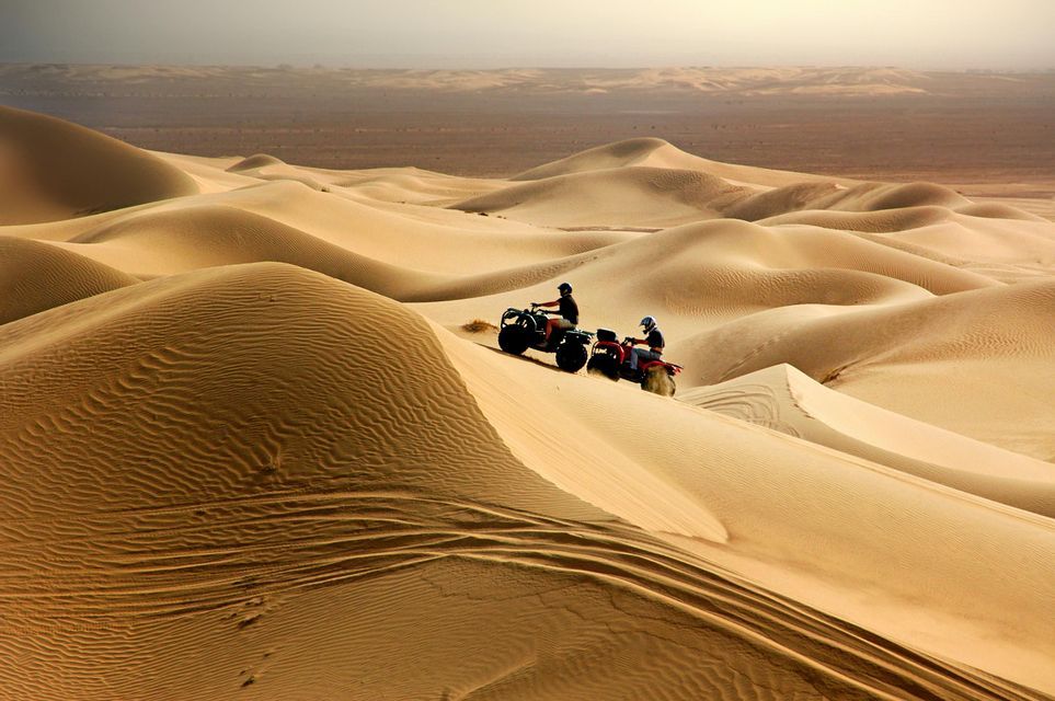 Un viaggio di gruppo WeRoad di due persone a bordo di quad lungo il fianco di una grande duna di sabbia in un vasto deserto.