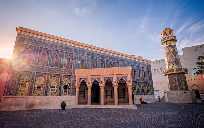 Una moschea ornata con piastrelle a mosaico blu e oro e un minareto si erge in un cortile acciottolato al tramonto