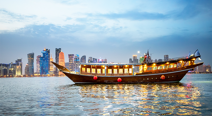Un tradizionale dhow in legno, illuminato dall'interno, galleggia sull'acqua al tramonto con lo skyline di una città moderna sullo sfondo.