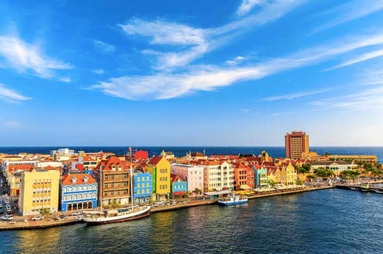 Un paesaggio urbano di edifici colorati lungo un lungomare, con barche ormeggiate nel porto e l'oceano sullo sfondo.