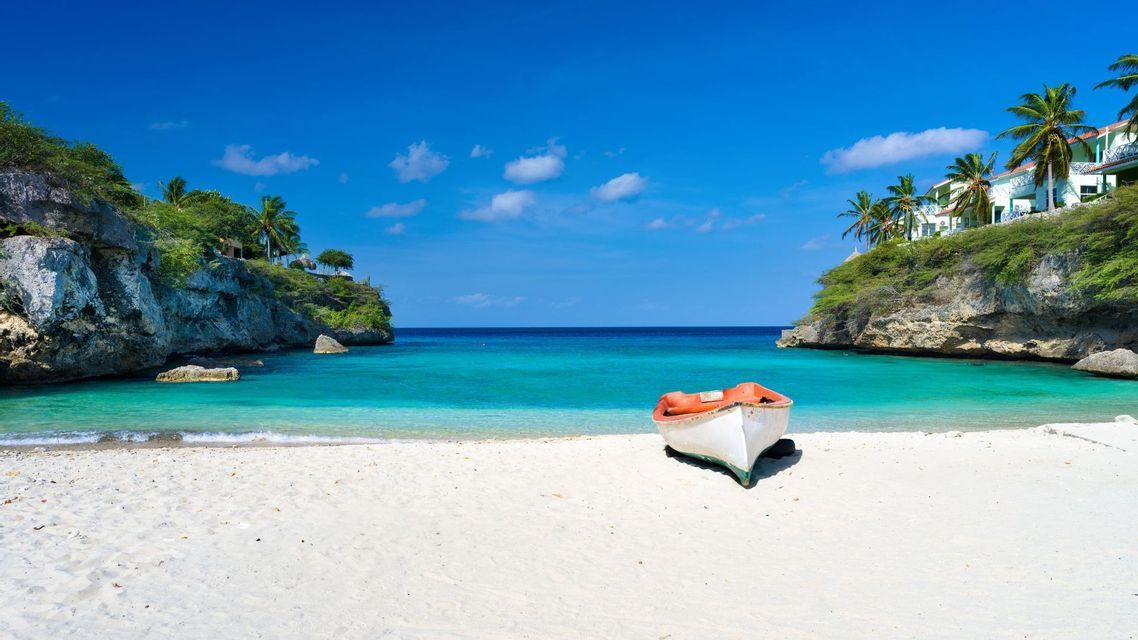 Una piccola barca con interni arancioni riposa su una spiaggia di sabbia bianca in una cala con acqua turchese e scogliere coperte di vegetazione.