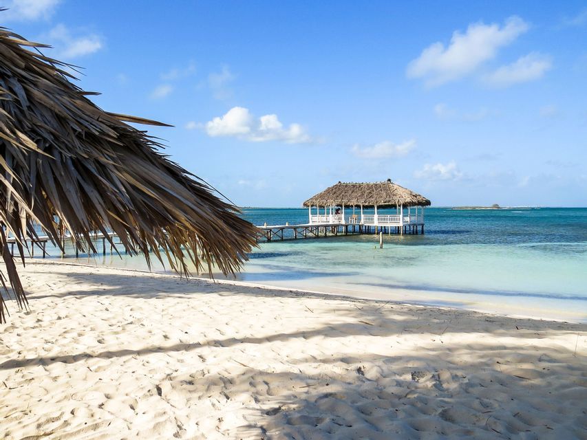 Un gazebo con tetto di paglia su un molo si estende su acque oceaniche turchesi da una spiaggia di sabbia bianca sotto un cielo blu.
