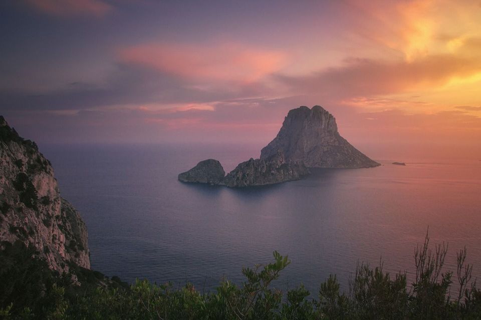 Una gran isla rocosa emerge de un mar en calma, vista desde un acantilado bajo un cielo de atardecer rosado y naranja.