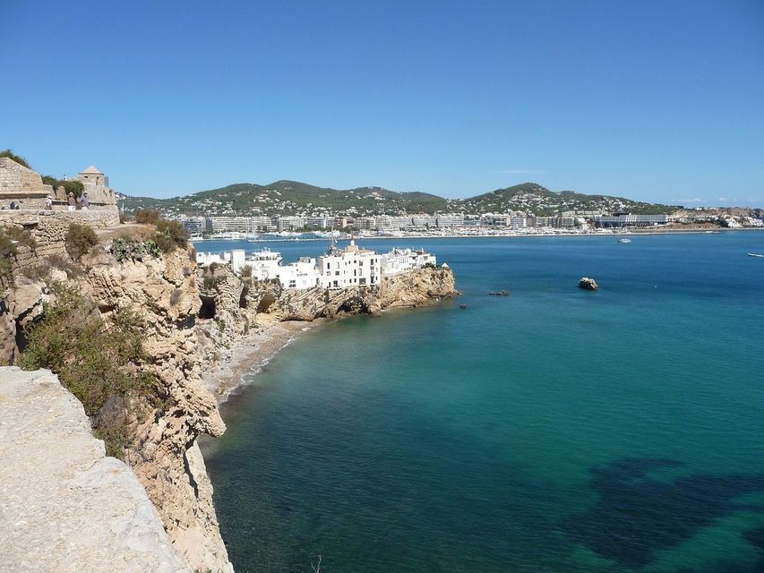 Una vista desde un acantilado de edificios blancos en una costa rocosa con vistas a una bahía turquesa bajo un cielo azul claro.